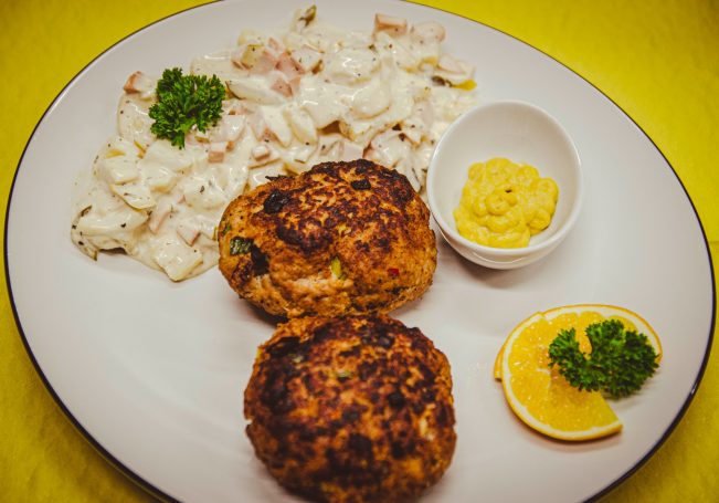 Zwei goldbraune Fischfrikadellen mit Remoulade, Salat und Zitronenscheiben auf einem Teller.