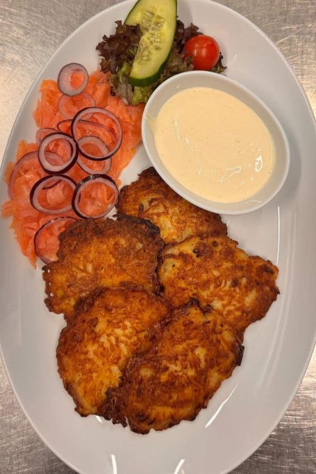 Kross gebratene Kartoffelpuffer mit Lachs, Salat, Zwiebeln und einer kleinen Schale Sauce.